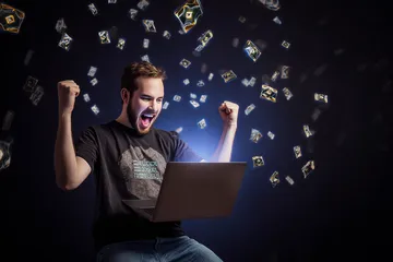 A man celebrating a winning moment on his laptop while gaming symbols float around him, capturing the excitement of KKFB games.